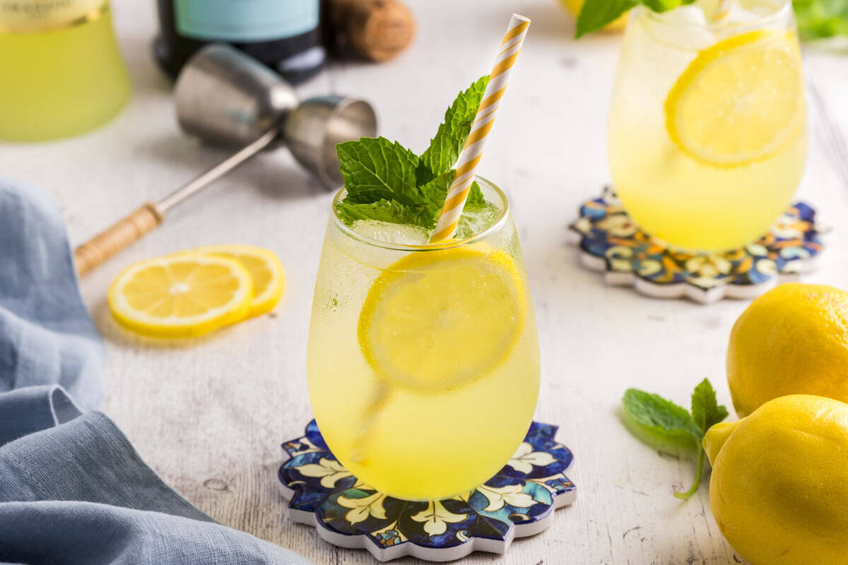 Side shot of a lemon spritz served in a non-stemmed wine glass. A lemon wheel is floating inside and up against the wall of the glass filled with ice. And a sprig of mint sits on top. A yellow and white straw is ready for sipping.