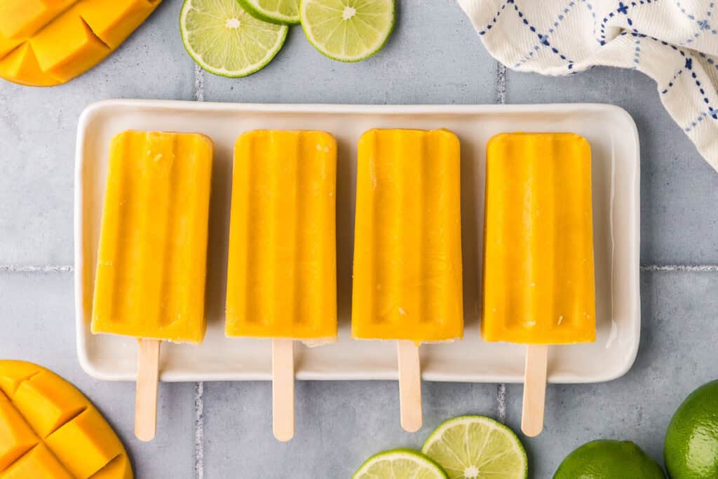 overhead shot of 4 bright orange mango popsicles laying side by side on a narrow white platter.
