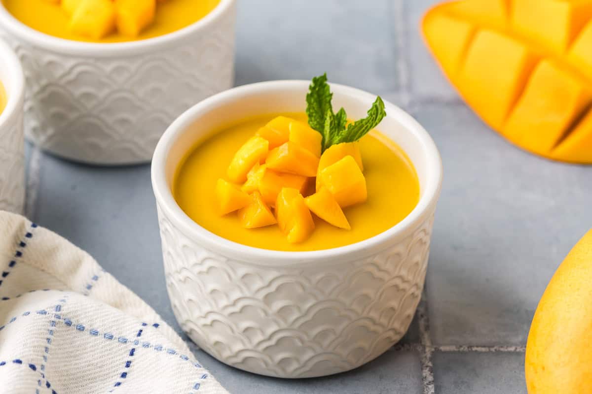 A 45 degree angle shot of a white ramekin filled with mango pudding. On top of the pudding is some diced fresh mango a sprig of fresh mint.