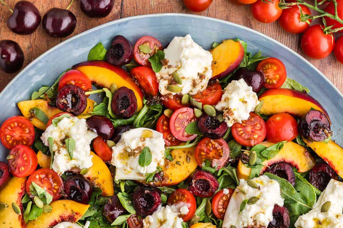 overhead shot of a peach burrata salad coming in to the scene from the bottom. At the top are fresh cherries and cherry tomatoes.