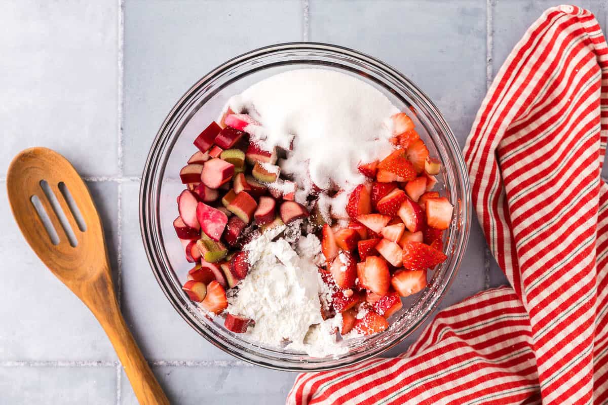 Overhead shot of a glass mixing bowl filled with chopped rhubarb and strawberries, cornstarch, lemon juice, and white sugar.