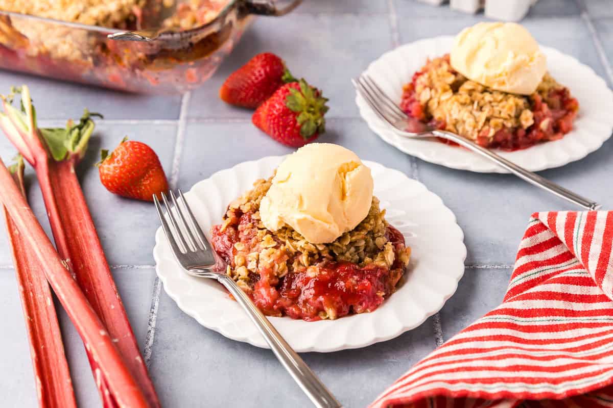 Side shot of a small white dessert plate filled with strawberry rhubarb crisp and a scoop of vanilla ice cream on top. Some rhubarb starks and strawberries are arranged near the plate. Another plate of the strawberry rhubarb crisp is in the right hand top corner.