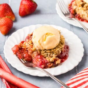 Side shot of a white dessert plate of strawberry rhubarb crisp and a fork. There is a scoop of vanilla ice cream on top.