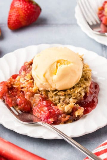 Side shot of a white dessert plate of strawberry rhubarb crisp and a fork. There is a scoop of vanilla ice cream on top.