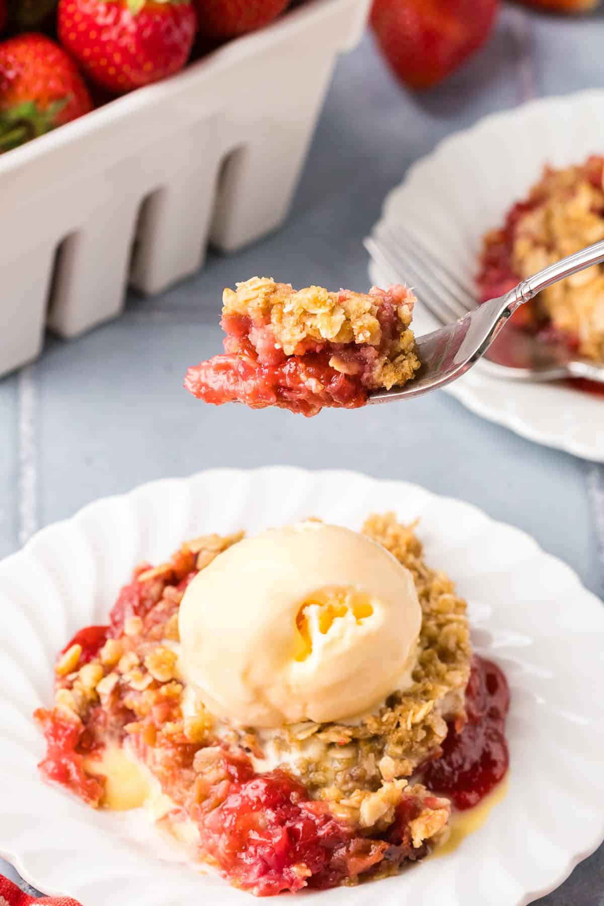 Side angle of a bite shot of strawberry rhubarb crisp. A fork is lifting up a portion of the crisp showing off it's pink jammy bottom and crispy oaty top.