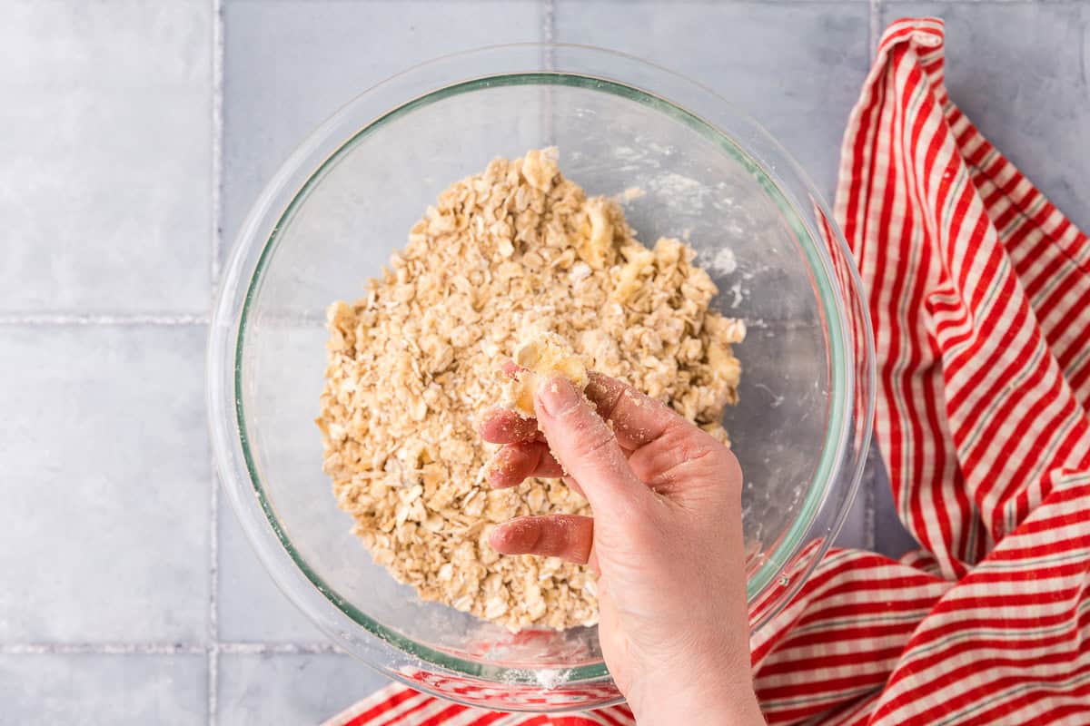overhead shot of all the crisp ingredients for the strawberry rhubarb crisp in a glass bowl. A hand is showing how to squish the butter into the dry ingredients.
