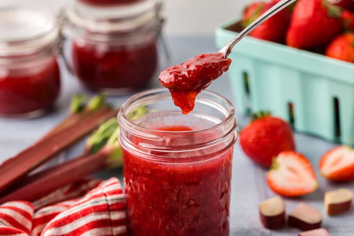 horizontal side shot of a jam jar filled with red strawberry rhubarb jam. A small silver spoon is scooping a heap of the jam out. In the background are strawberries and fresh rhubarb.