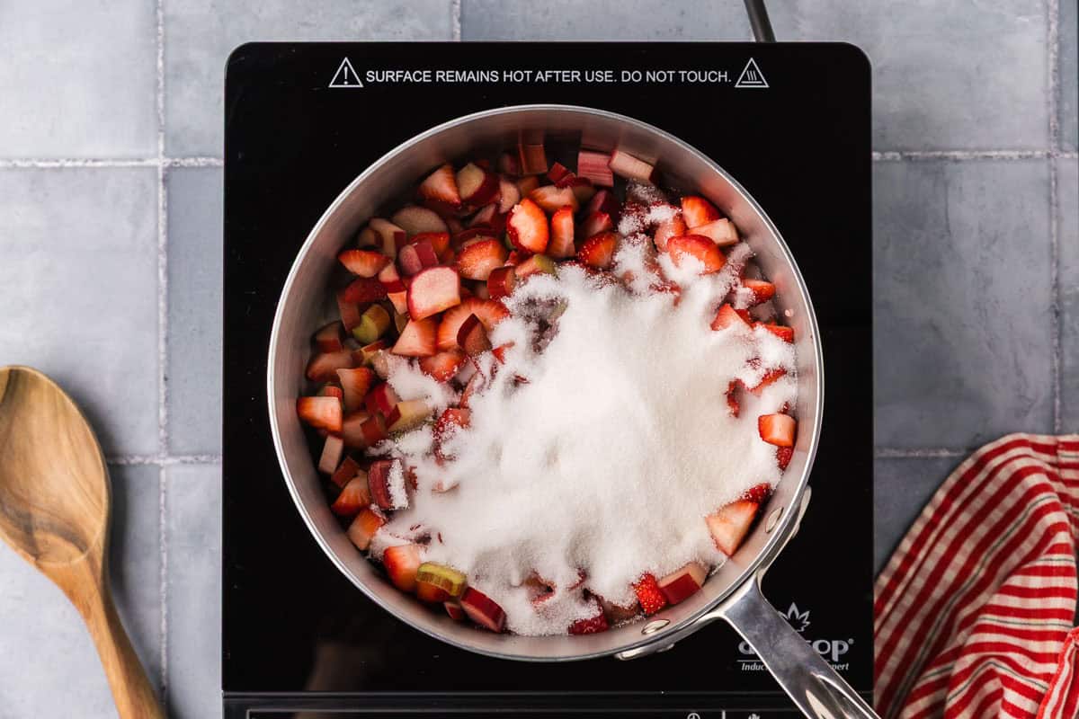 overhead shot of a saucepan filled with strawberries, rhubarb, white sugar, and lemon juice.