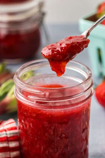 Side shot of strawberry rhubarb jam being scooped out of a jam jar with a small silver spoon. In the background are strawberries and fresh rhubarb.