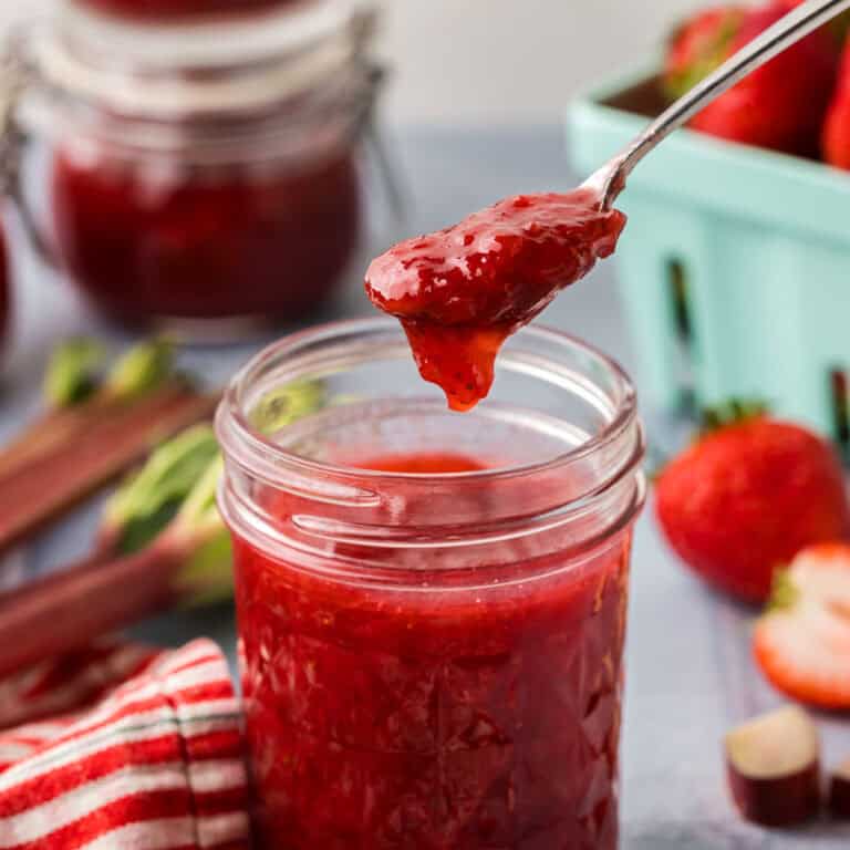 Side shot of strawberry rhubarb jam being scooped out of a jam jar with a small silver spoon. In the background are strawberries and fresh rhubarb.