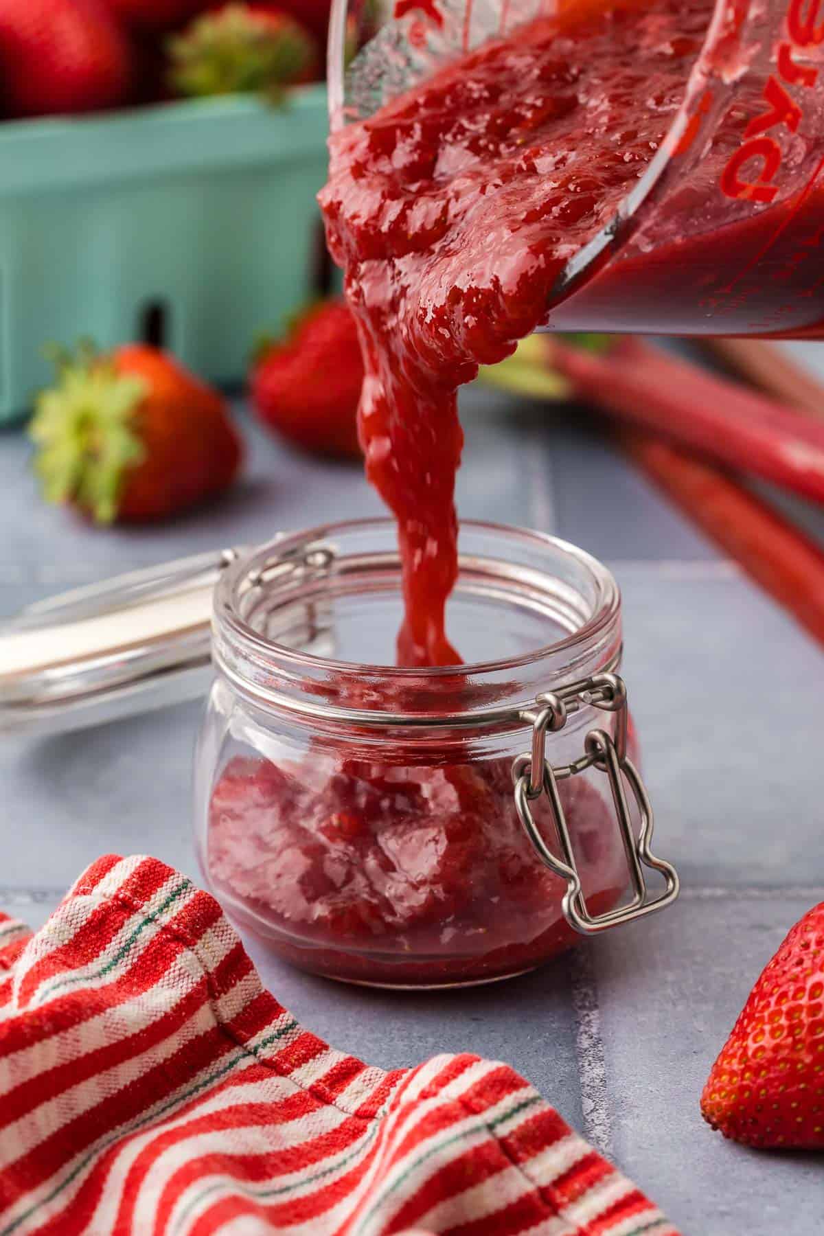 side shot of the thick strawberry rhubarb jam being poured into jam jars for storage. The jam is thick and red.