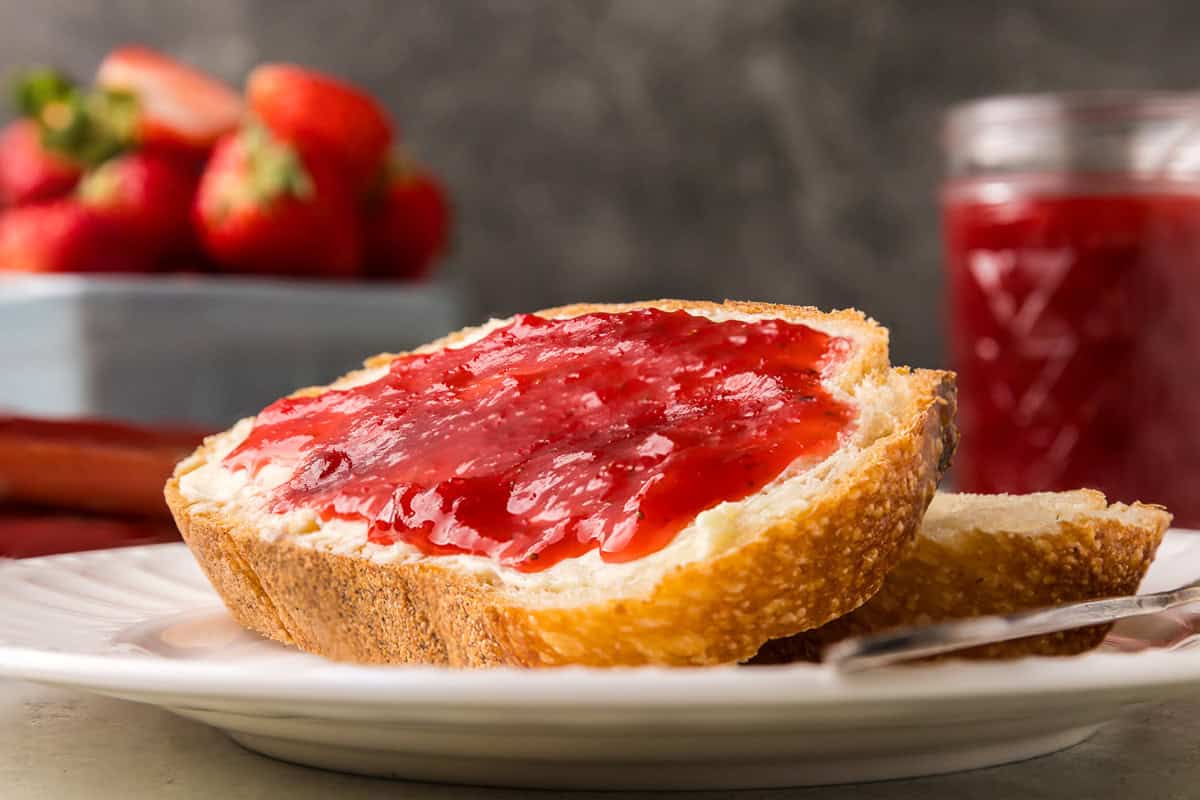 Very close up shot of a piece of sourdough toast smothered with butter and strawberry rhubarb jam. The jam is a glossy pink/red and is thick but not chunky.