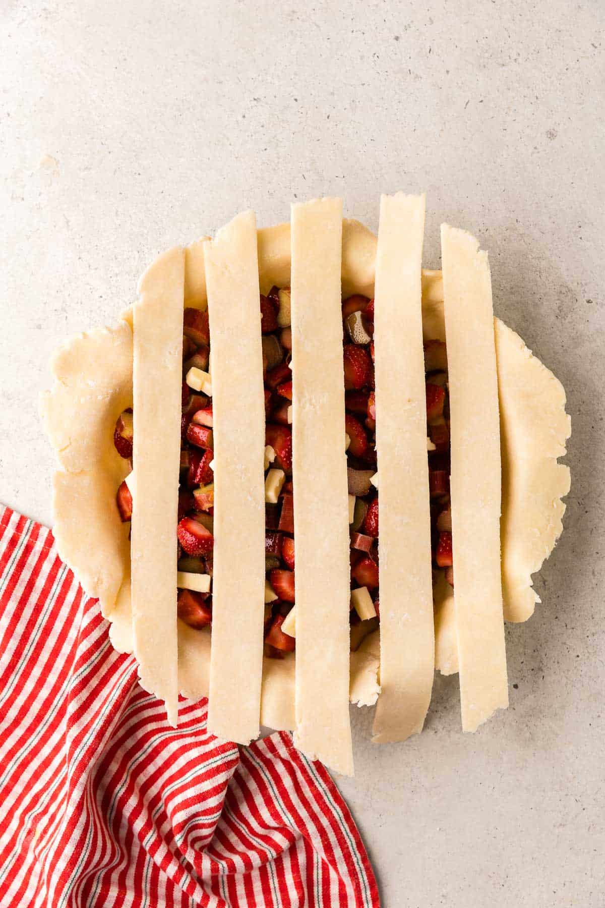 Overhead shot of a pie shell filled with strawberry rhubarb pie filling, and five strips of dough layed across vertically with gaps between each strip.