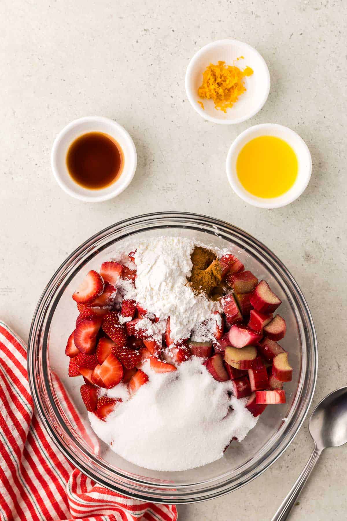 An overhead shot of a glass mixing bowl with the strawberries, rhubarb, sugar, cornstarch, and cinnamon added. Above the bowl the following ingredients are waiting to be added: vanilla extract, orange zest, and orange juice.