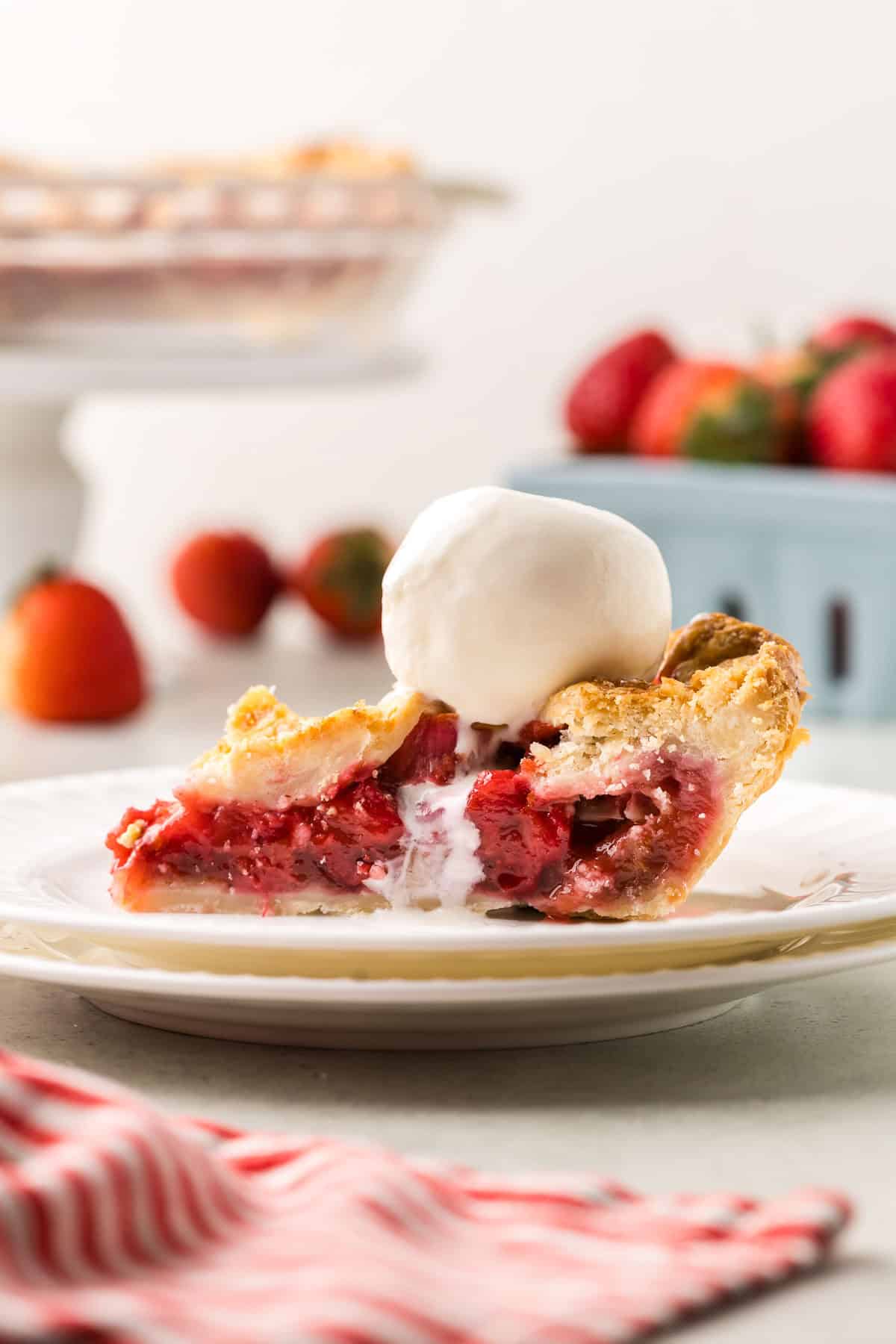 side shot of a slice of strawberry rhubarb pie with a scoop of vanilla ice cream melting on the top.