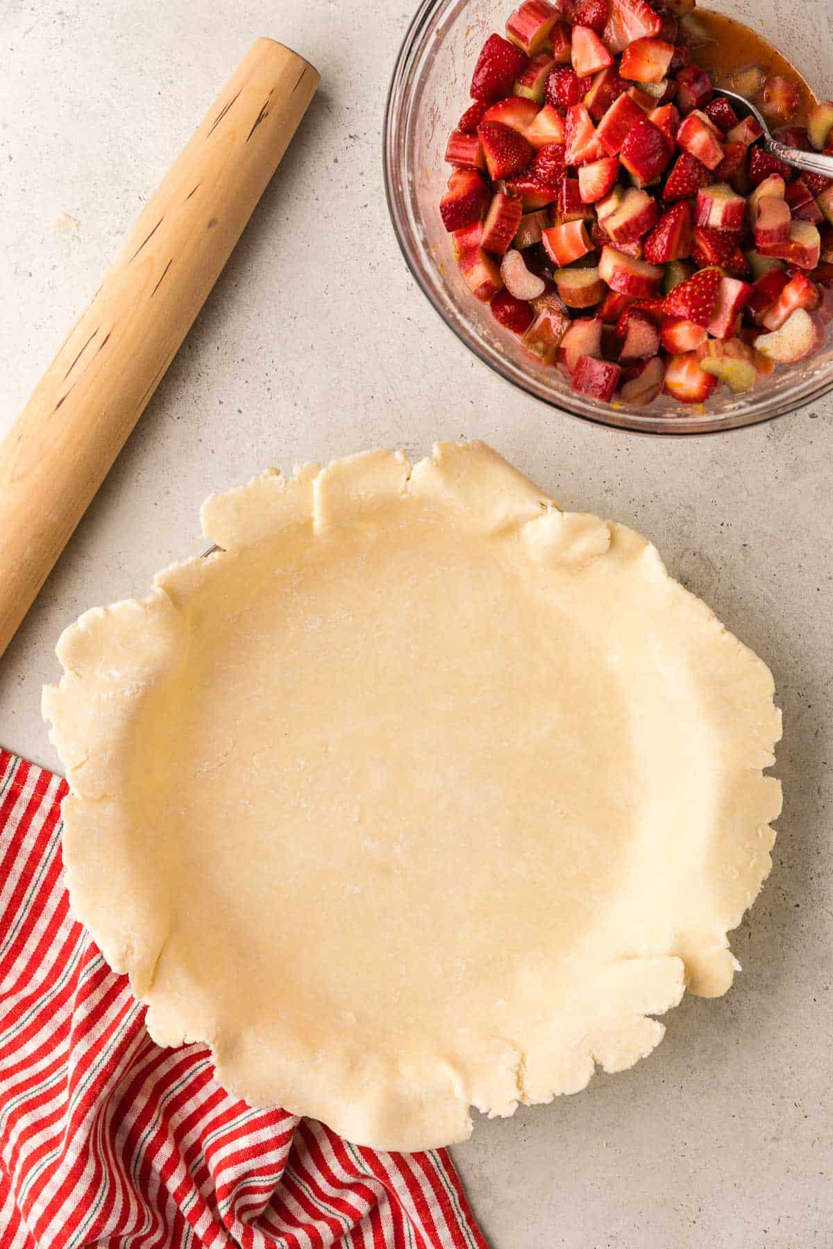 Overhead shot of pie dough that has been rolled out and seated into a pie dish. Above the pie is a bowl with the strawberry rhubarb filling.