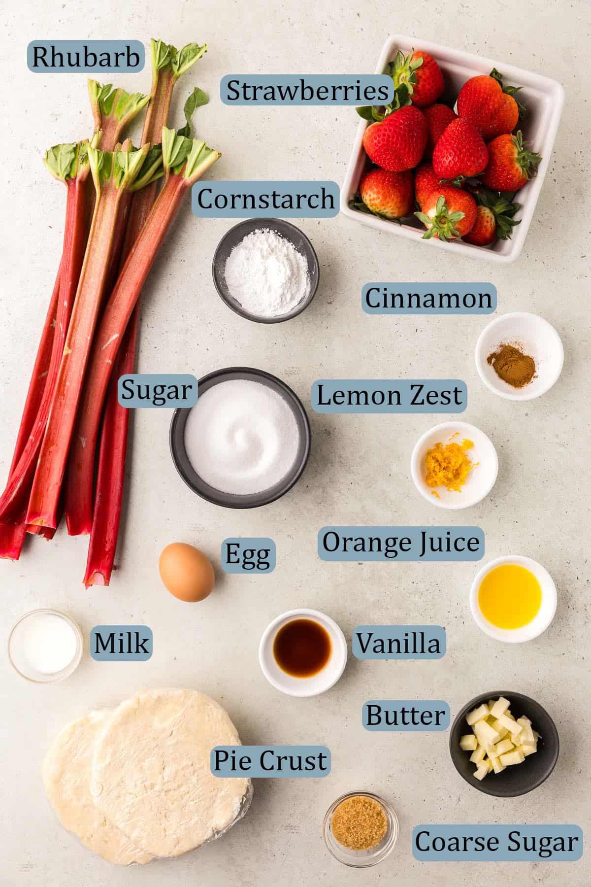 overhead shot of all the ingredients needed to make a strawberry rhubarb pie. Ingredients are: fresh rhubarb and strawberries, cornstarch, granulated sugar, cinnamon, orange zest, orange juice, egg, milk, butter, vanilla extract, coarse sugar, and double pie crust dough.