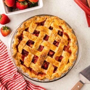 Overhead shot of a strawberry rhubarb pie with a lattice top in a glass pie dish. Peeking-in in the corners are fresh strawberries and rhubarb.