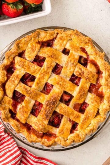 Overhead shot of a strawberry rhubarb pie with a lattice top in a glass pie dish. Peeking-in in the corners are fresh strawberries and rhubarb.