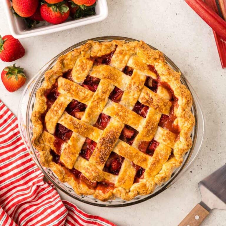 Overhead shot of a strawberry rhubarb pie with a lattice top in a glass pie dish. Peeking-in in the corners are fresh strawberries and rhubarb.