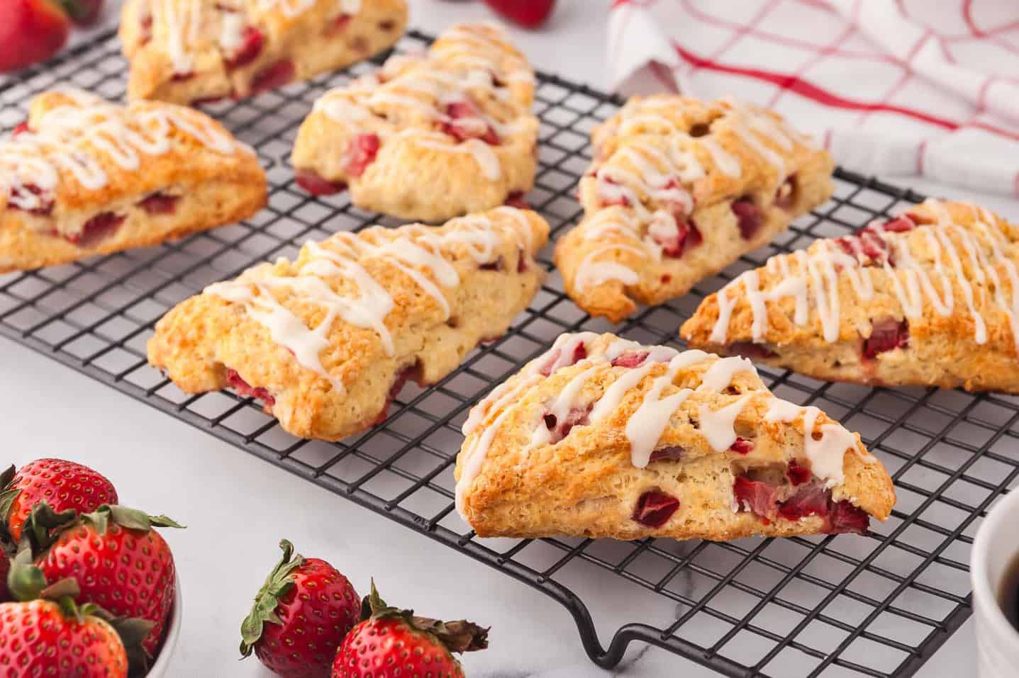 A side shot of strawberry scones cooling on a cooling rack. Each scone has a glaze drizzled across, and fresh strawberries are in the background and foreground.