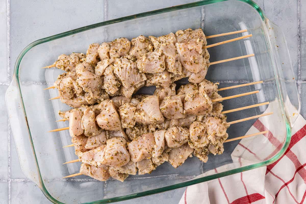 overhead shot of a glass casserole dish filled with the skewered marinated chicken. Each skewer is filled with chicken and lots of oregano can be seen.