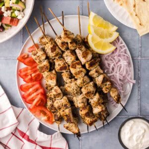 Overhead shot of a white serving plate filled with skewers of chicken souvlaki garnished with sliced tomatoes, red onions, and wedges of fresh lemon.