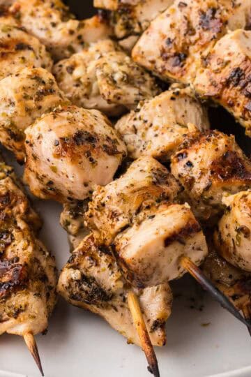 Close-up shot of chicken souvlaki skewers. Each one has grill marks and is glistening with marinade and flecks of oregano.