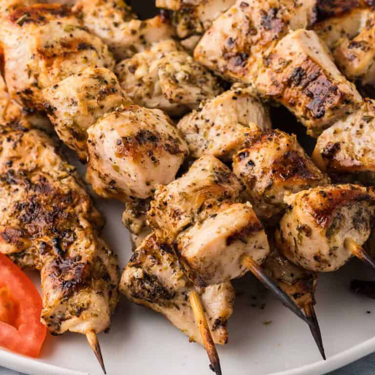 Close-up shot of chicken souvlaki skewers. Each one has grill marks and is glistening with marinade and flecks of oregano.