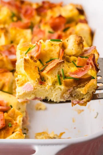 Side shot looking at a slice of eggs benedict casserole being removed from the casserole dish with a metal spatula. The square slice is made with chunks of English muffin, and Canadian ham, held together with custardy egg. Chives have been sprinkled on top.