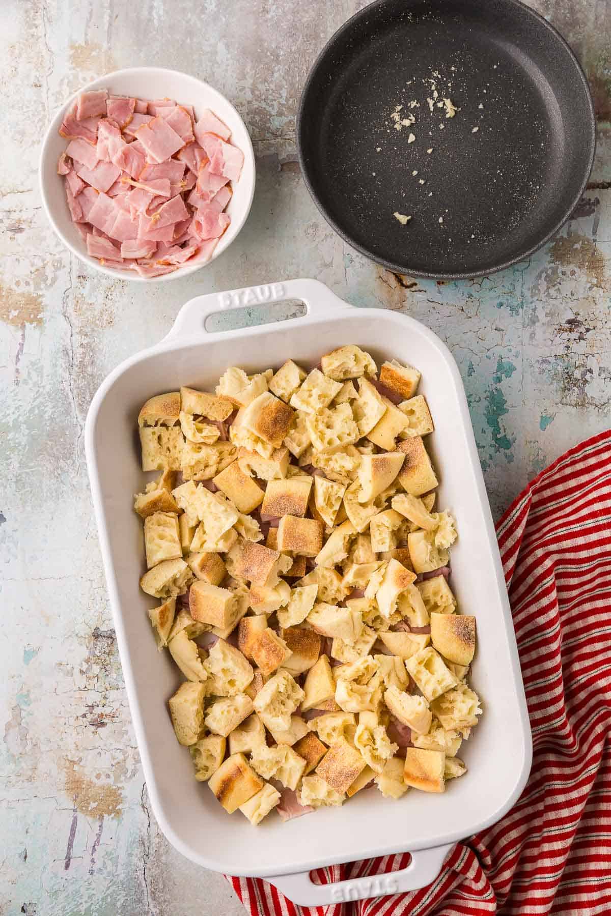 A white 9x13 casserole dish with a layer of torn pieces of English muffin added on top of the back bacon.