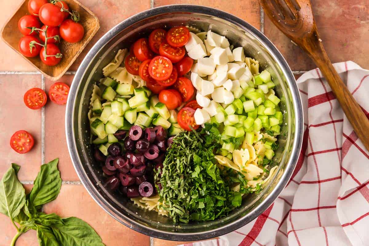 Overhead shot looking in to a large metal mixing bowl. All the ingredients going in to the pasta salad are added in a pie shape on top. There is halved cherry tomatoes, bocconcini, celery, fresh basil and parsley, kalamata olive, and cucumbers.