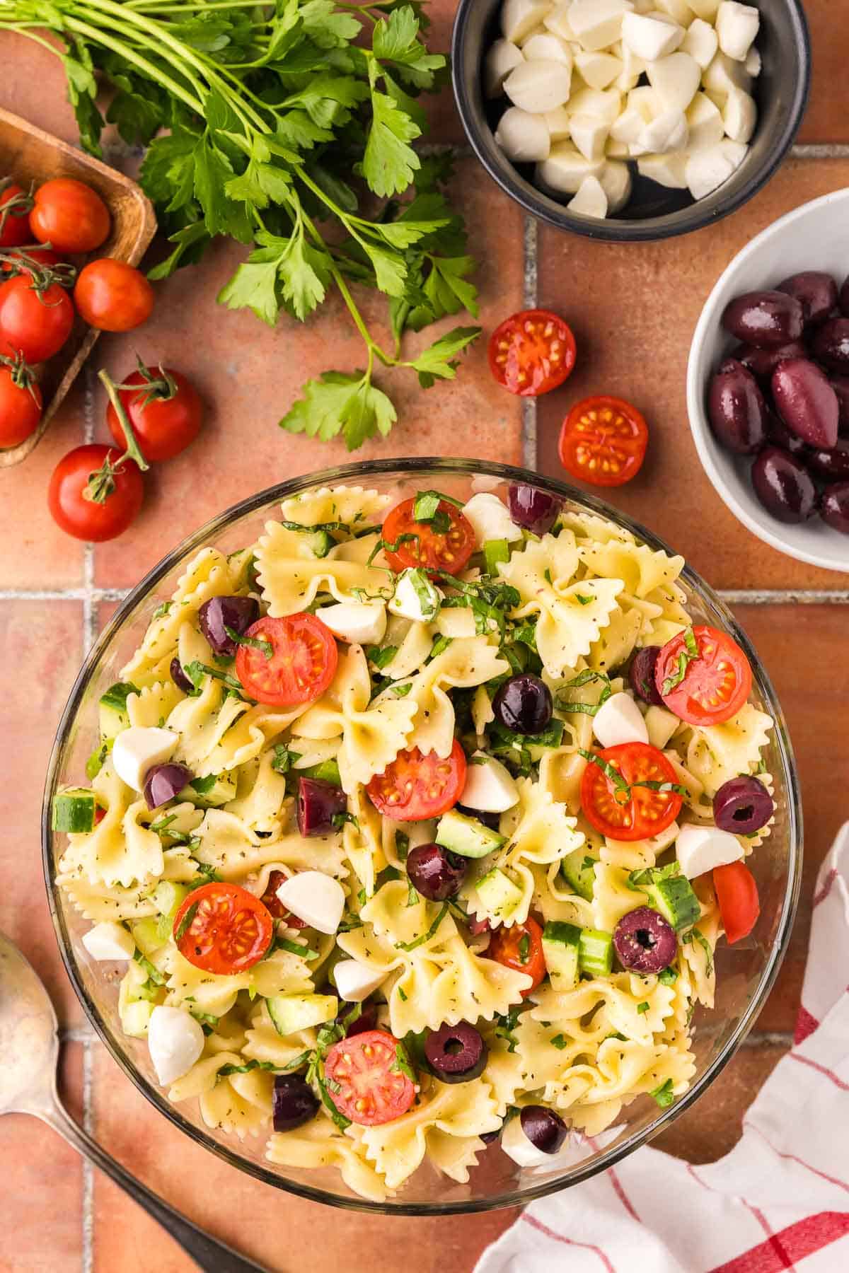 overhead shot of a bowl filled with pasta salad with Italian flavors. The noodles are bow tie shaped and lots of cherry tomatoes, celery, cucumber, kalamata olives and bocconcini are around.