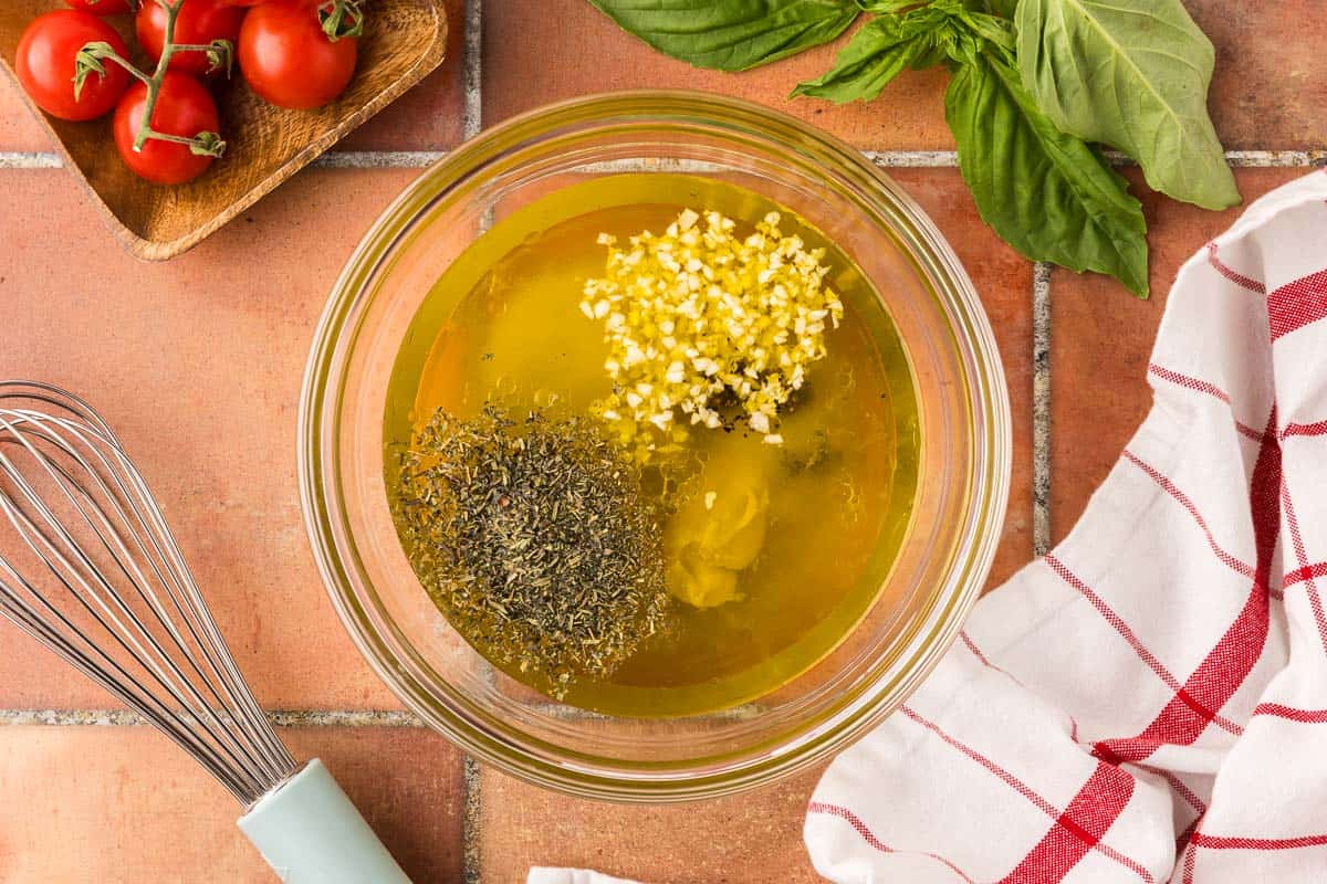 Overhead shot looking into a glass mixing bowl filled with the ingredients for the vinaigrette. There is olive oil, apple cider vinegar, lemon juice, Dijon mustard, sugar, salt, and floating on top is freshly chopped garlic, Italian seasoning, and black pepper.