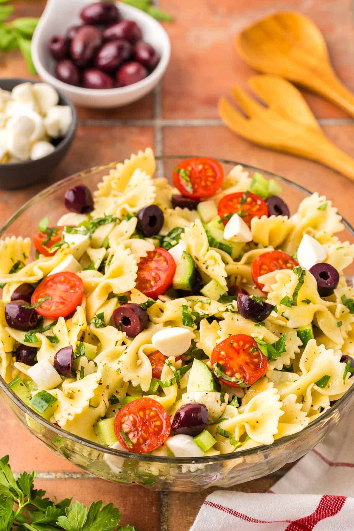 Side shot of a glass bowl filled with bow tie Italian Pasta Salad. The bow tie noodles are surrounded by halved cherry tomatoes, chopped cucumbers, celery, kalamata olives and bocconcini.