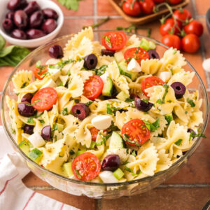Side shot of a glass bowl filled with Italian Pasta Salad. Bow tie pasta is surrounded by halved cherry tomatoes, chopped cucumbers, celery, kalamata olives and bocconcini. In the background is a bowl of kalamata olives and fresh cherry tomatoes on the vine.