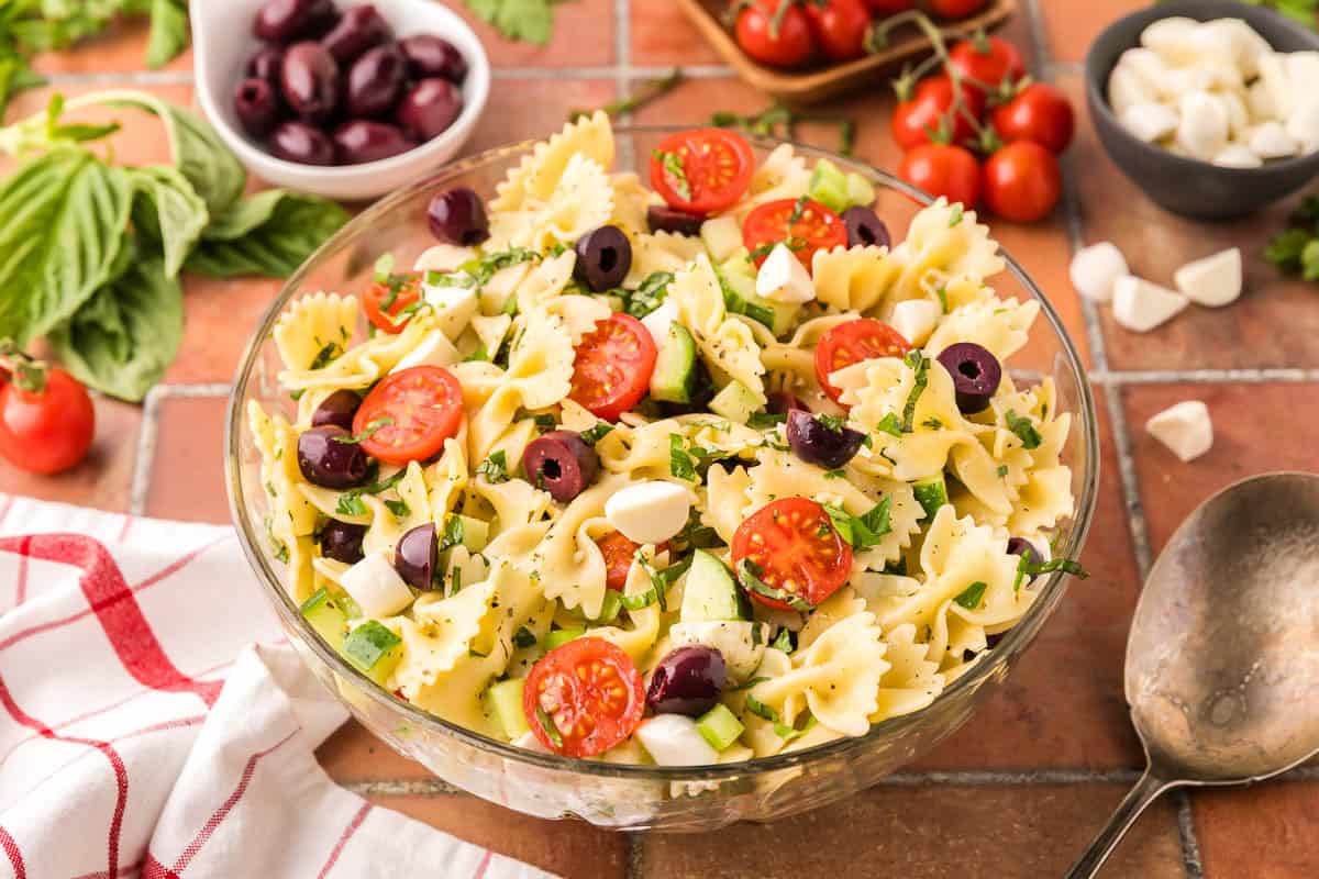 Side shot of a glass bowl filled with bow tie Italian Pasta Salad. The bow tie noodles are surrounded by halved cherry tomatoes, chopped cucumbers, celery, kalamata olives and bocconcini. In the background is fresh basil, kalamata olives, cherry tomatoes, and bocconcini.