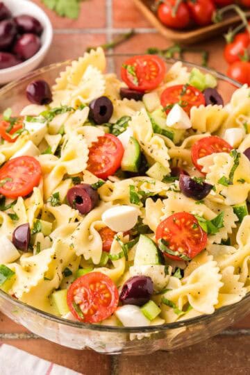 Side shot of a glass bowl filled with bow tie Italian Pasta Salad. The bow tie noodles are surrounded by halved cherry tomatoes, chopped cucumbers, celery, kalamata olives and bocconcini. In the background is fresh basil, kalamata olives, cherry tomatoes, and bocconcini.