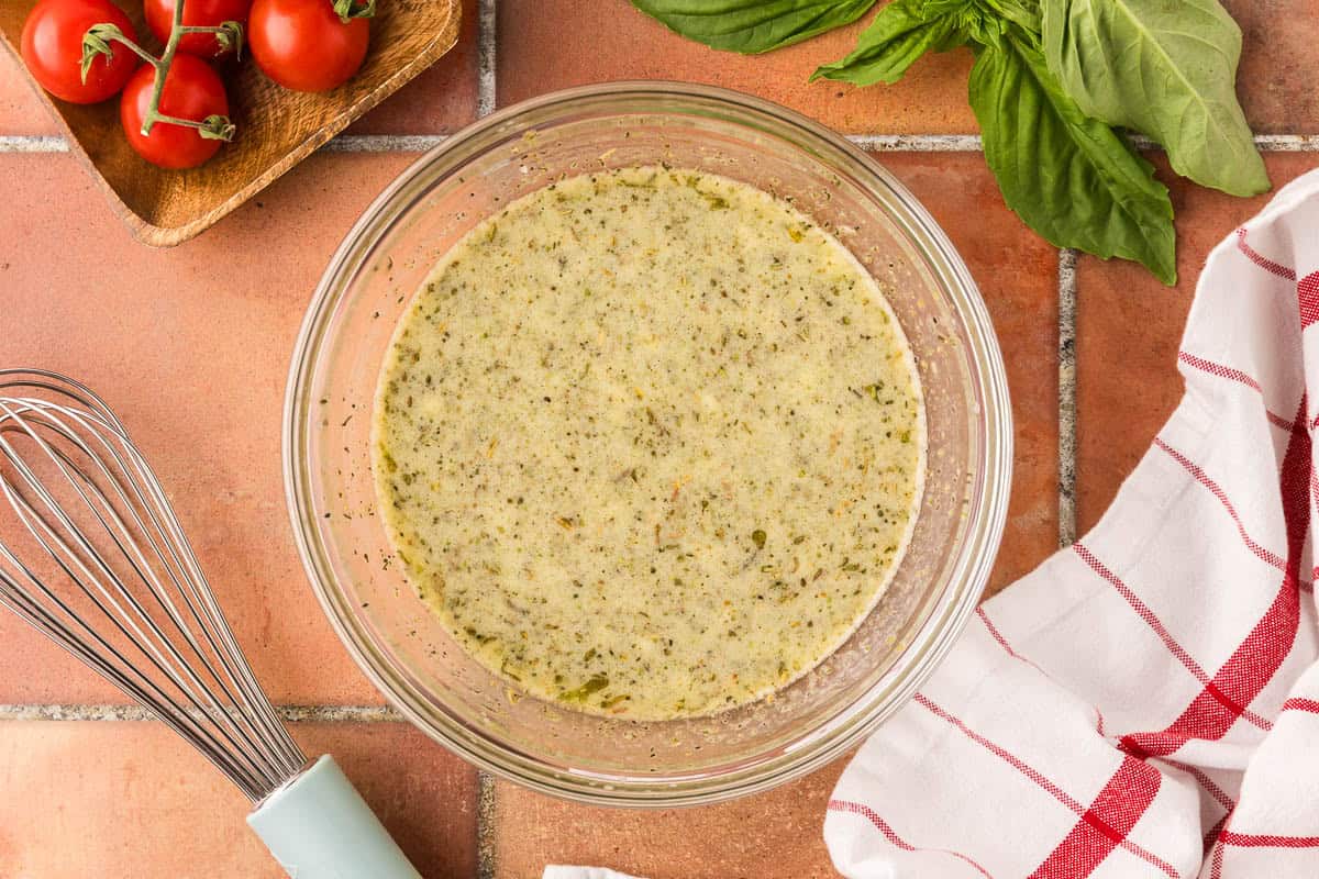 overhead shot of a glass bowl filled with the emulsified vinaigrette ingedients. It is a pale yellow color with seasonings floating on the surface.