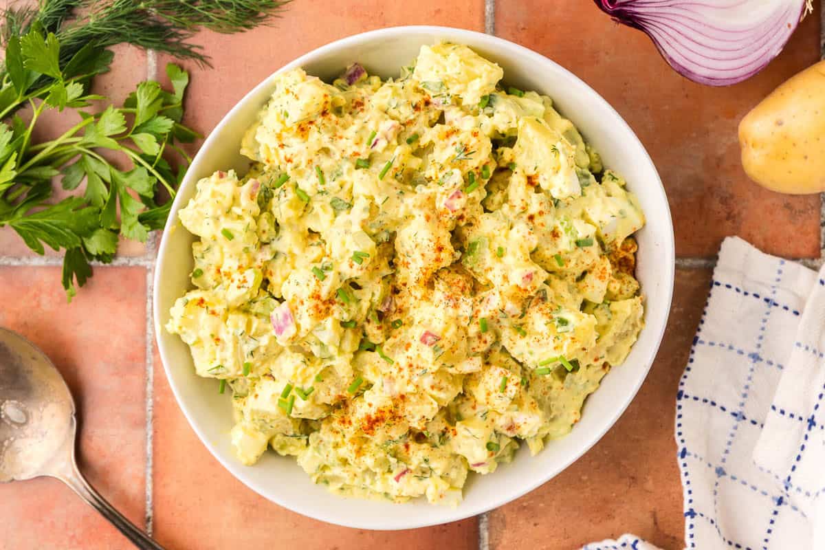 Overhead shot of a white ceramic bowl filled with a creamy yellow chunky potato salad. Fresh parsley, red onion are peeking into the scene.