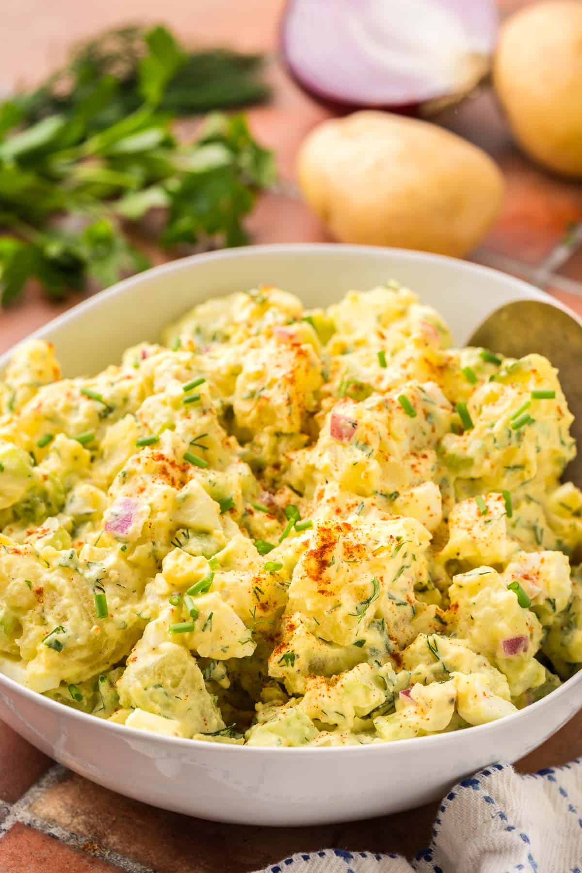 Side shot of a white ceramic bowl filled with a creamy yellow chunky potato salad. Fresh herbs, potatoes, and red onion are in the background.