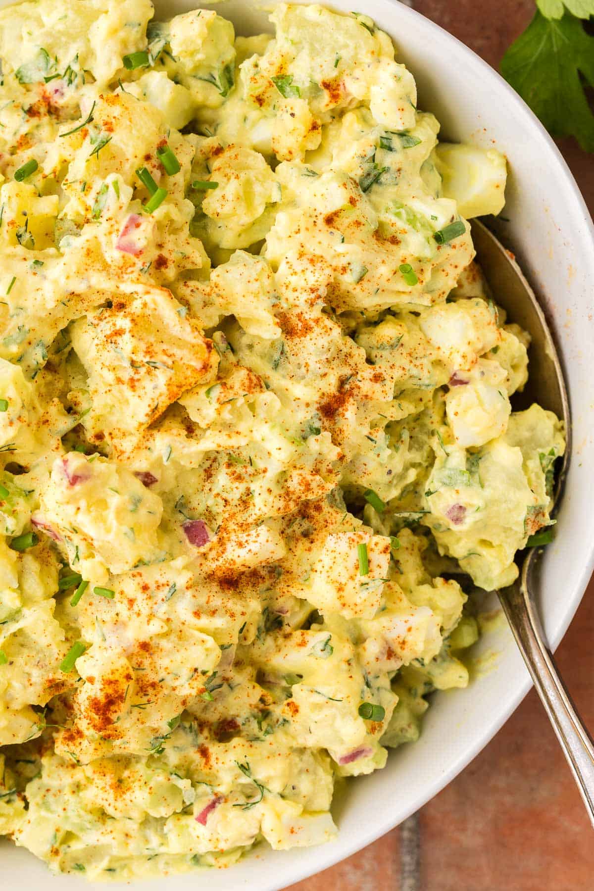 Overhead shot looking at a closeup of a bowl of creamy yellow potato salad which takes up most of the shot. The salad is a creamy yellow color and chunky with potatoes, celery and eggs. It is garnished with a sprinkle of paprika and some chopped chives.