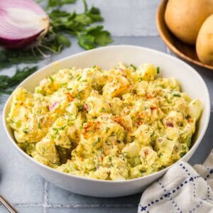 Side shot of a white ceramic bowl filled with a creamy yellow chunky potato salad. In the background is some red onion, fresh parsley and potatoes.