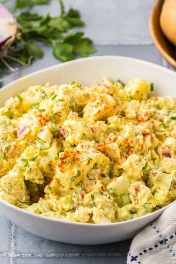 Side shot of a white ceramic bowl filled with a creamy yellow chunky potato salad. In the background is some red onion, fresh parsley and potatoes.