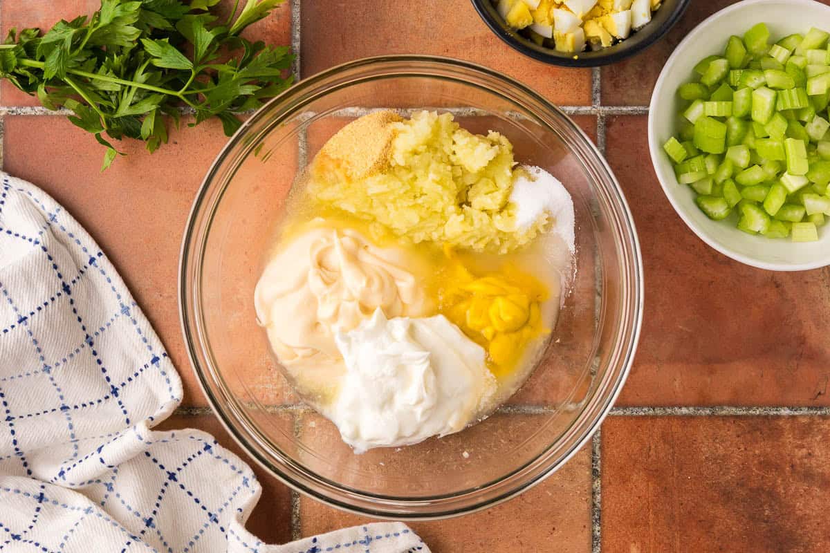 Overhead shot of a glass bowl filled with all the dressing ingredients. Present are: mayonnaise, Greek yogurt, yellow mustard, mashed potatoes, dill brine, and sugar.