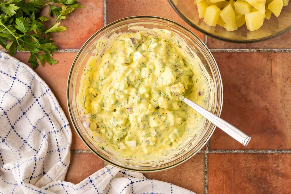 Overhead shot of the potato salad dressing bowl no also containing the celery, onion, dill pickles, fresh herbs, and boiled potatoes. It is a creamy yellow color and quite chunky.