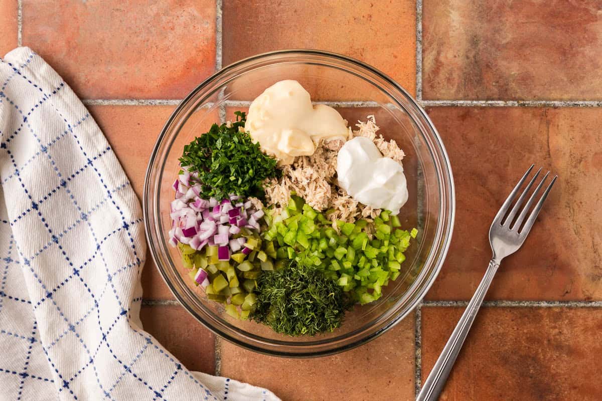 Overhead shot of a glass bowl filled with all the ingredients to make a tuna salad. There is tuna, mayo, Greek yogurt, chopped celery, red onion, dill pickle, fresh parsley and dill.