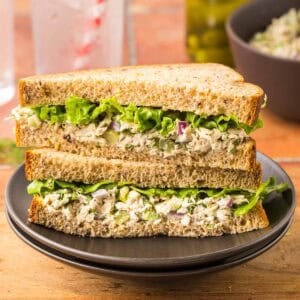 Side shot of a sandwich using fresh tuna salad. The bread is a whole wheat bread, and there is a fluted leafy piece of lettuce. Sandwich has been cut in half on the diagonal and the two slices stacked together revealing the creamy tuna salad and leafy lettuce.