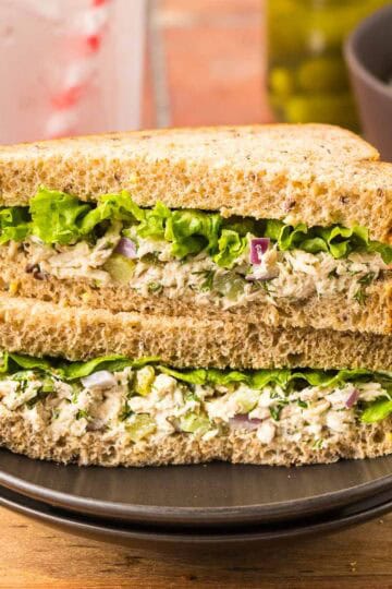 Side shot of a sandwich using fresh tuna salad. The bread is a whole wheat bread, and there is a fluted leafy piece of lettuce. Sandwich has been cut in half on the diagonal and the two slices stacked together revealing the creamy tuna salad and leafy lettuce.