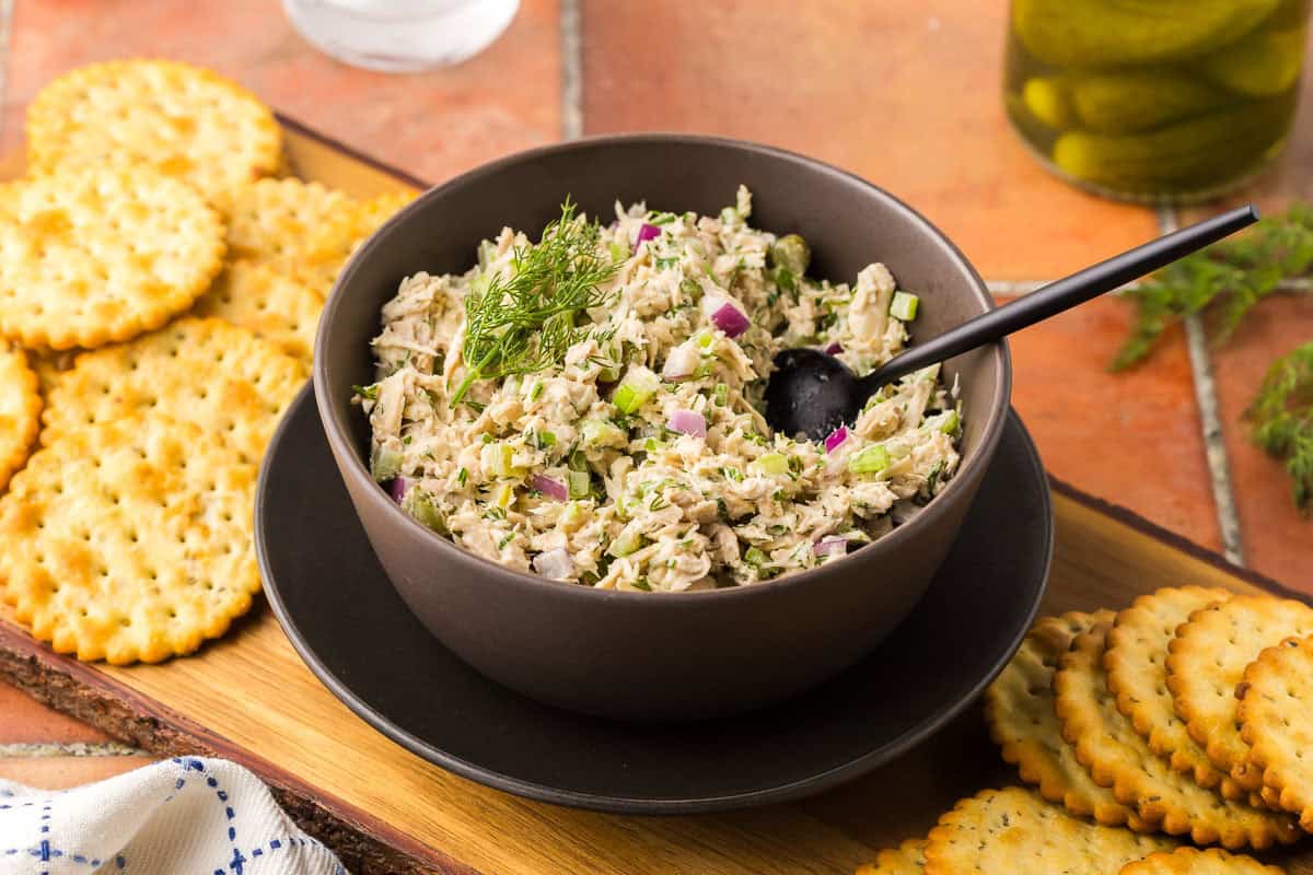 horizontal side shot of a small dark bowl filled with tuna salad. The bowl is sitting on a wooden cutting board surrounded by crackers. A small black serving spoon is resting in the salad.
